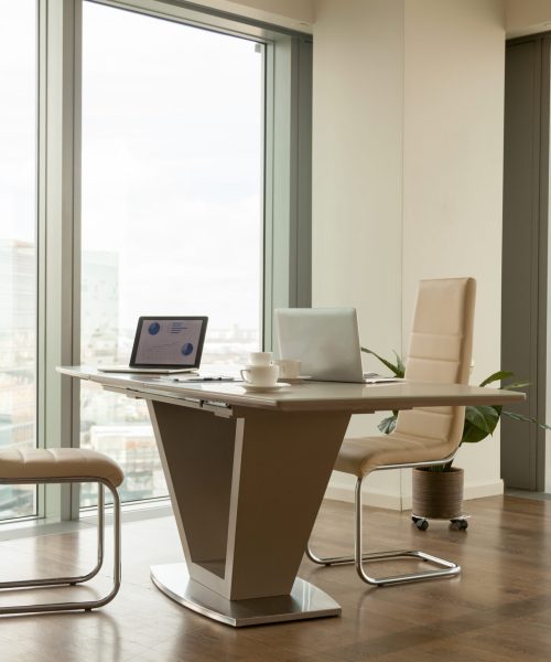 Image of work desk with two laptops on it and comfortable armchairs beside in bright office interior with large windows and urban landscape outside. Modern office workplace, negotiation room design